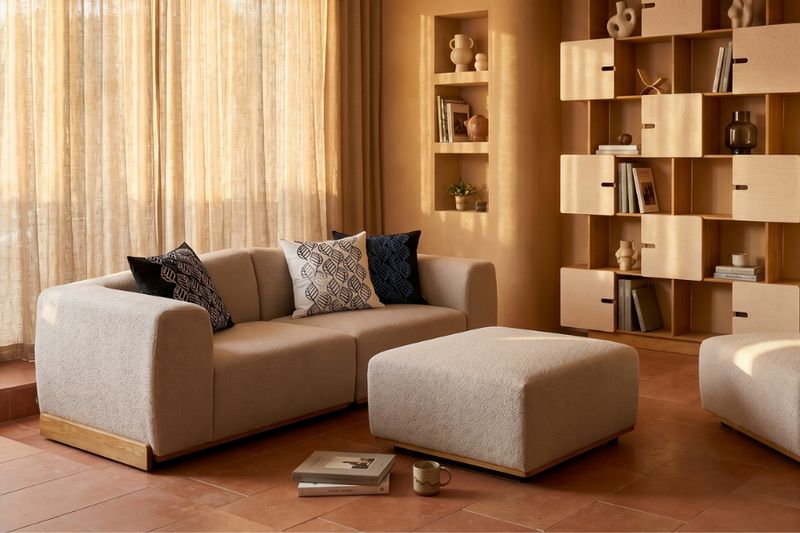 Sofa SAILER in Sandbeige mit Holzsockel und gemusterten Kissen vor einem offenen Regal, Hocker davor