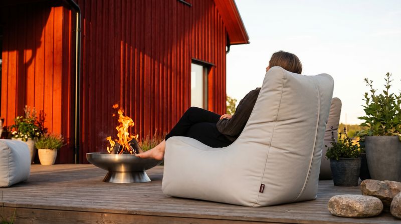 Person sitzt in einem Sitzsack-Sessel auf einer Holzterrasse vor einem roten Holzhaus, daneben eine Feuerschale
