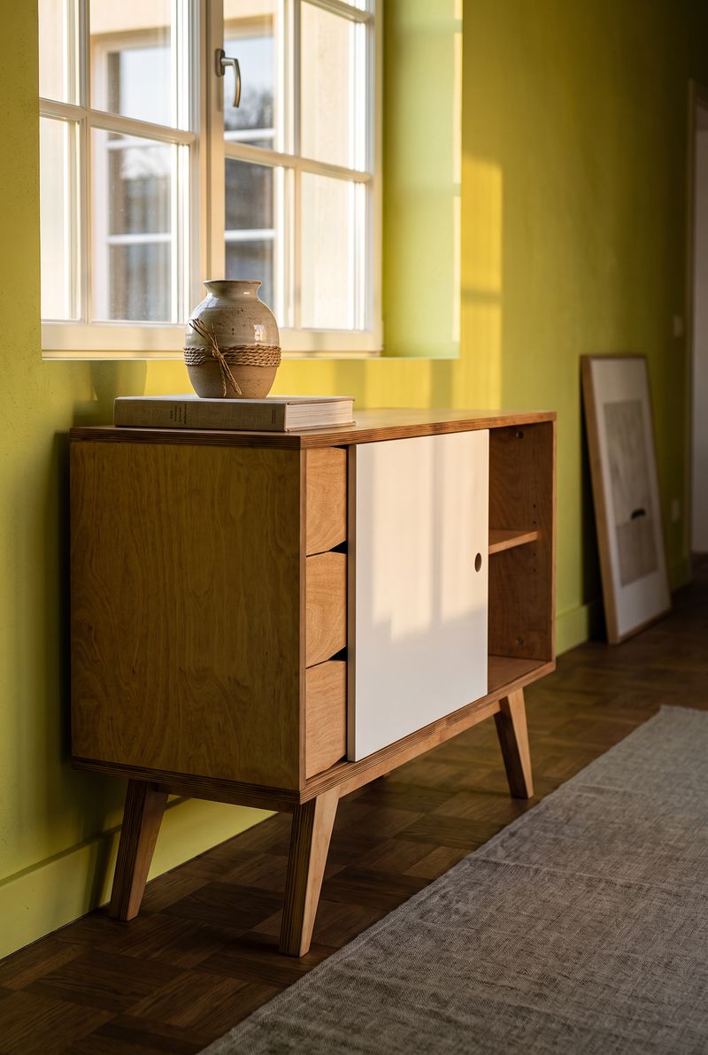 Sideboard aus Holz mit weisser Schiebetuer vor gelbgruener Wand, Keramikvase und Buecher darauf
