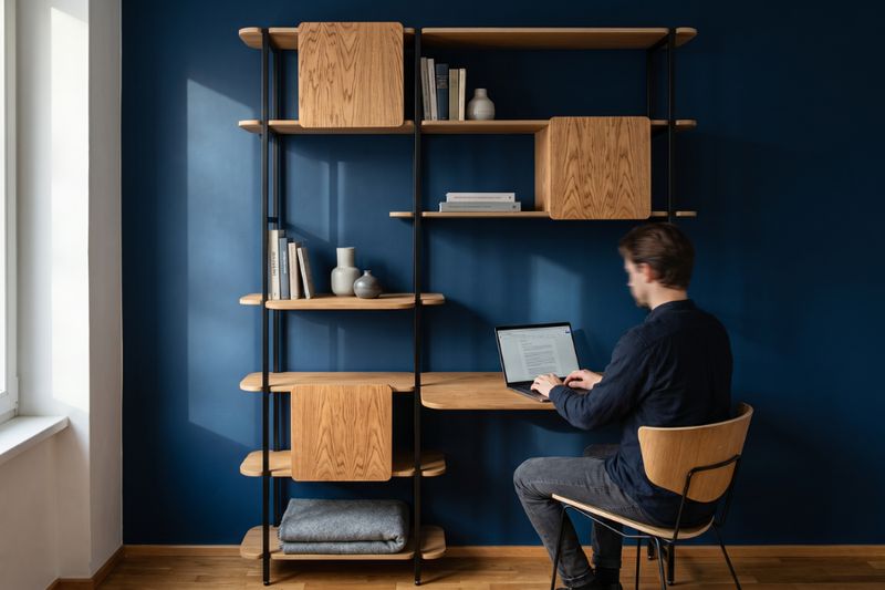 Mann arbeitet am Laptop an einem Wandregal aus Holz und Metall vor einer dunkelblauen Wand