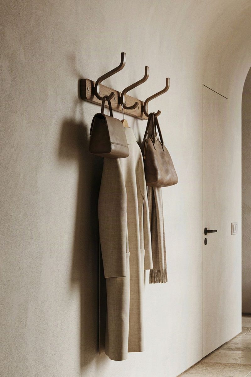 Wandgarderobe aus Holz mit drei Metallhaken, daran ein Mantel, eine Ledertasche und ein Schal, heller Putzwand-Flur